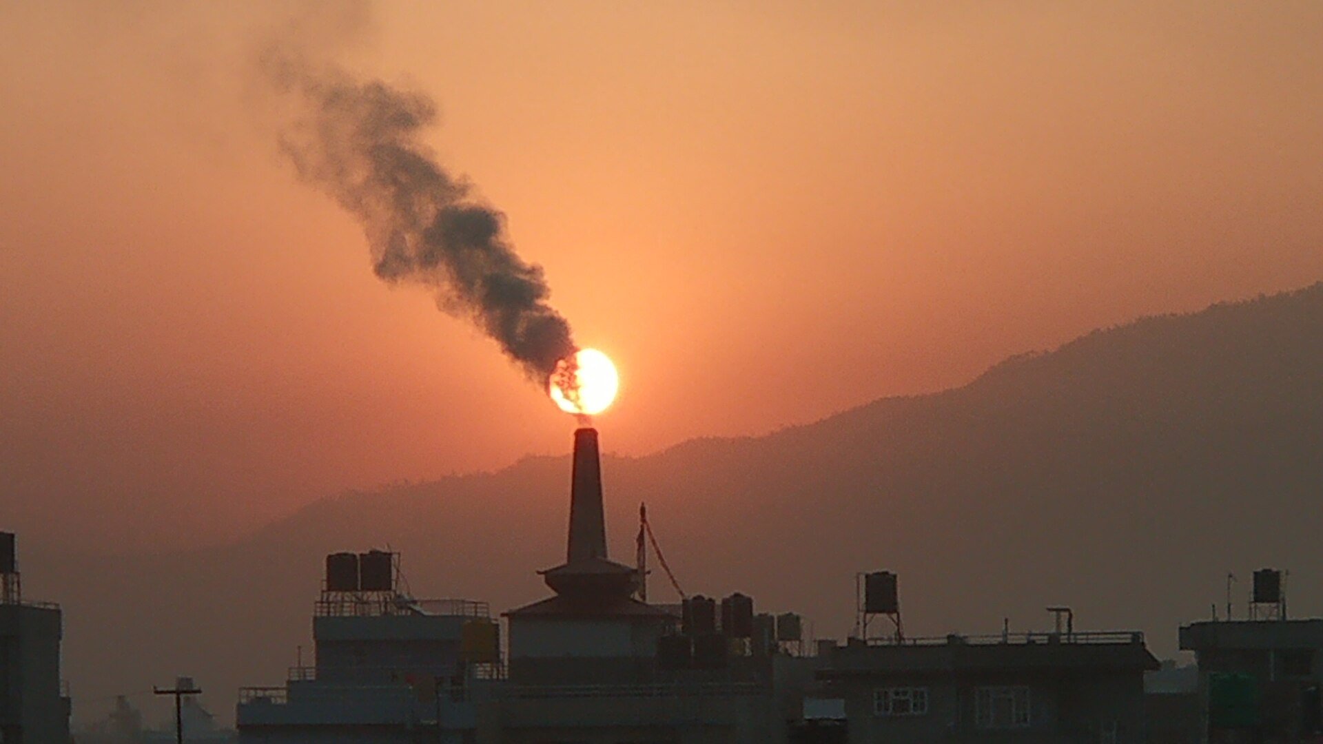 Industrial zone, smokestack with dark plume, sun centered behind stack, orange sunset glow, editorial travel photo, no people.