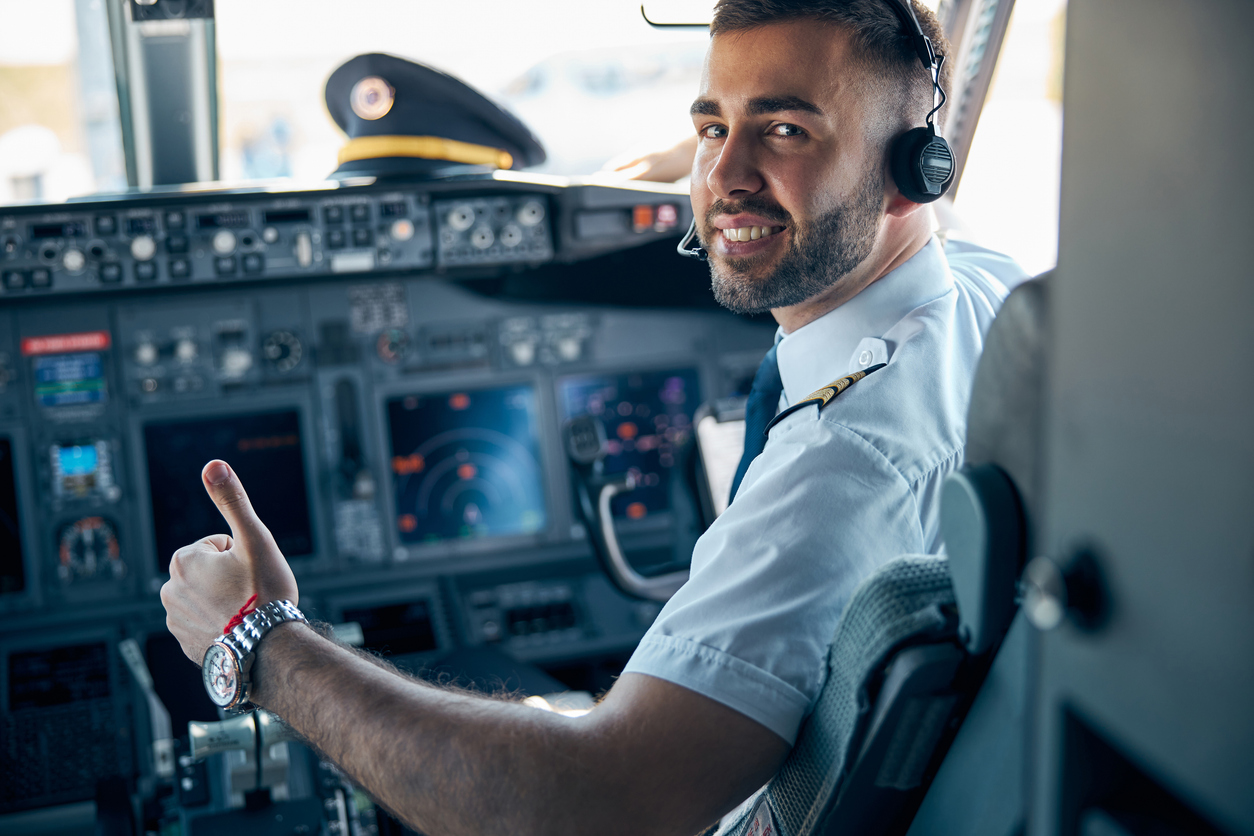 Airplane cockpit interior, pilot in headset thumbs-up, wide view with displays and captain’s hat, bright daylight, editorial travel photo, with person.