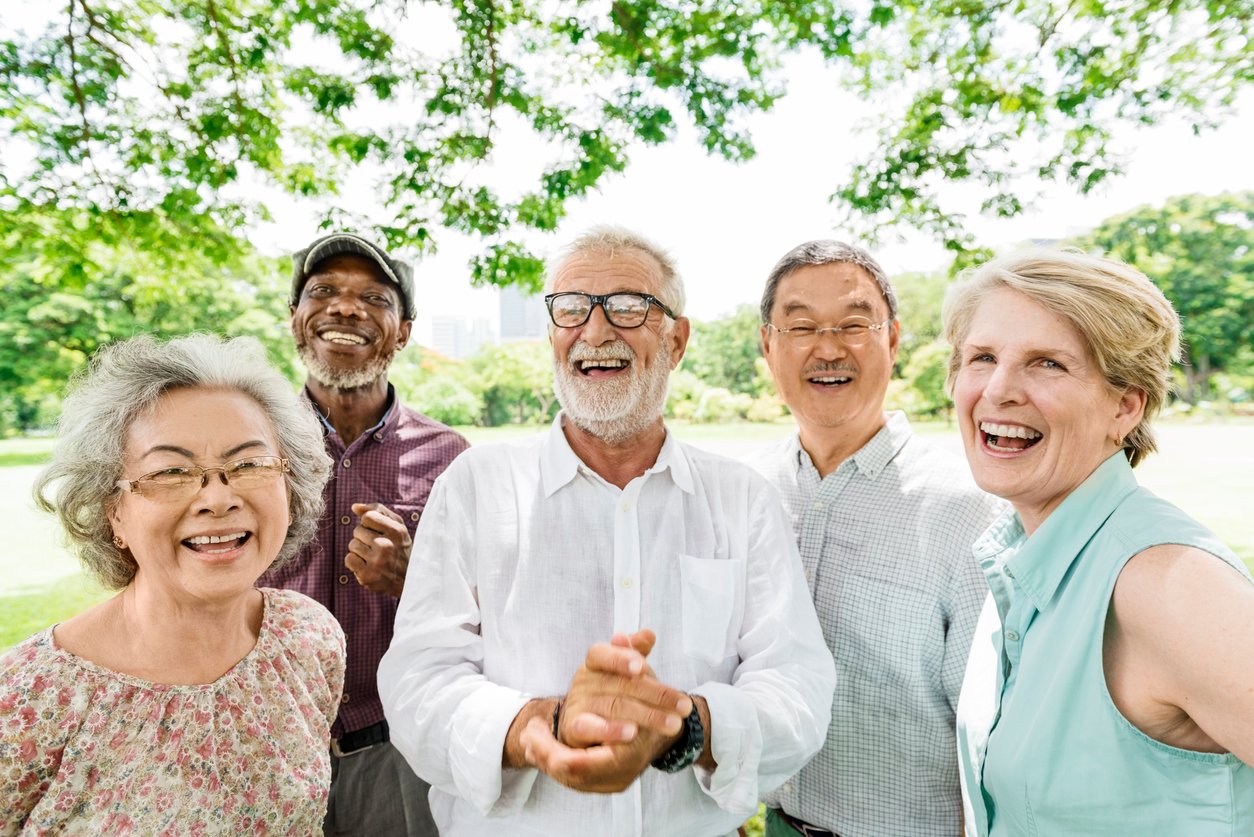 Park, five seniors standing close, group under leafy tree, bright sunlight, documentary style, people.