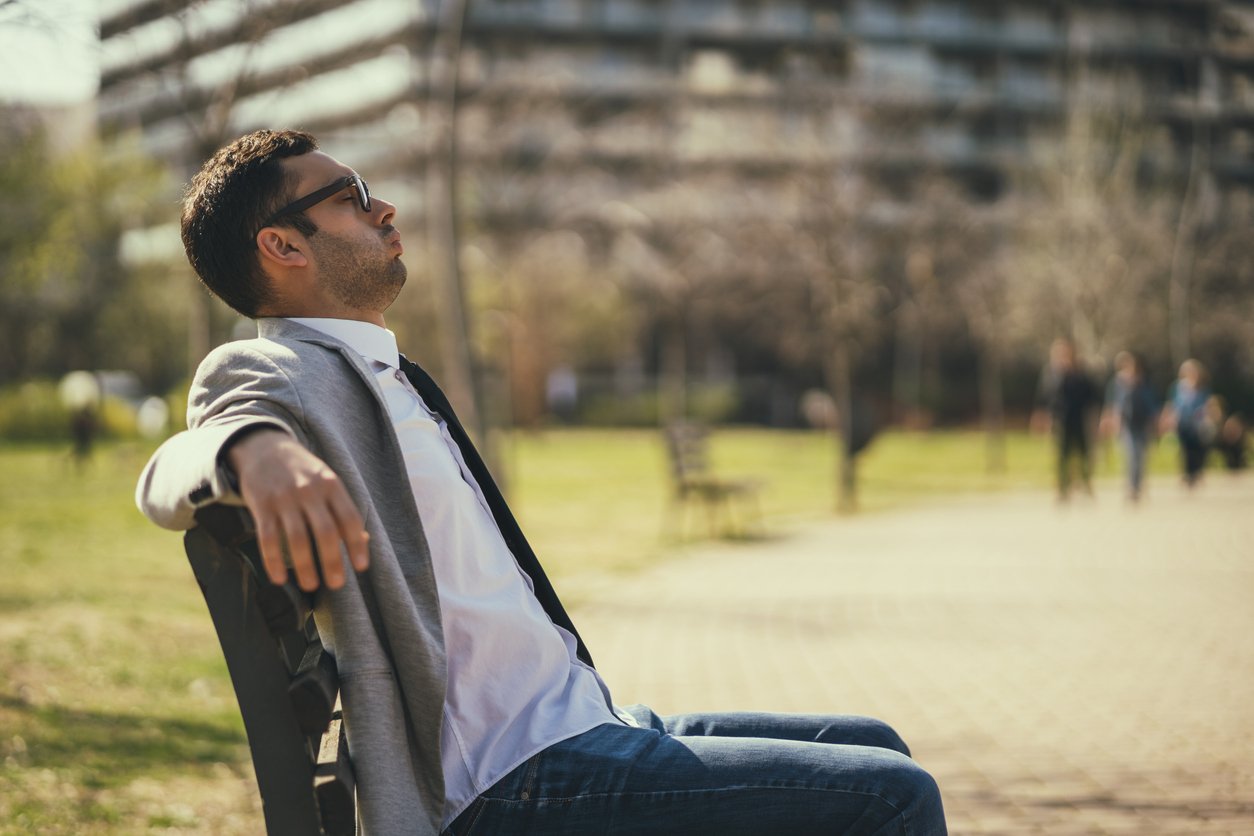 Park bench, man in sunglasses and blazer reclining, foreground focus, bright midday sun, editorial travel photo, people present.