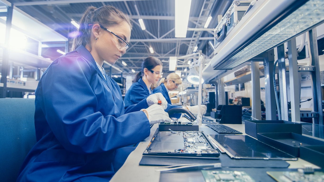 Modern factory floor, woman assembling electronics, workstation foreground, bright artificial lights, documentary style, three people.