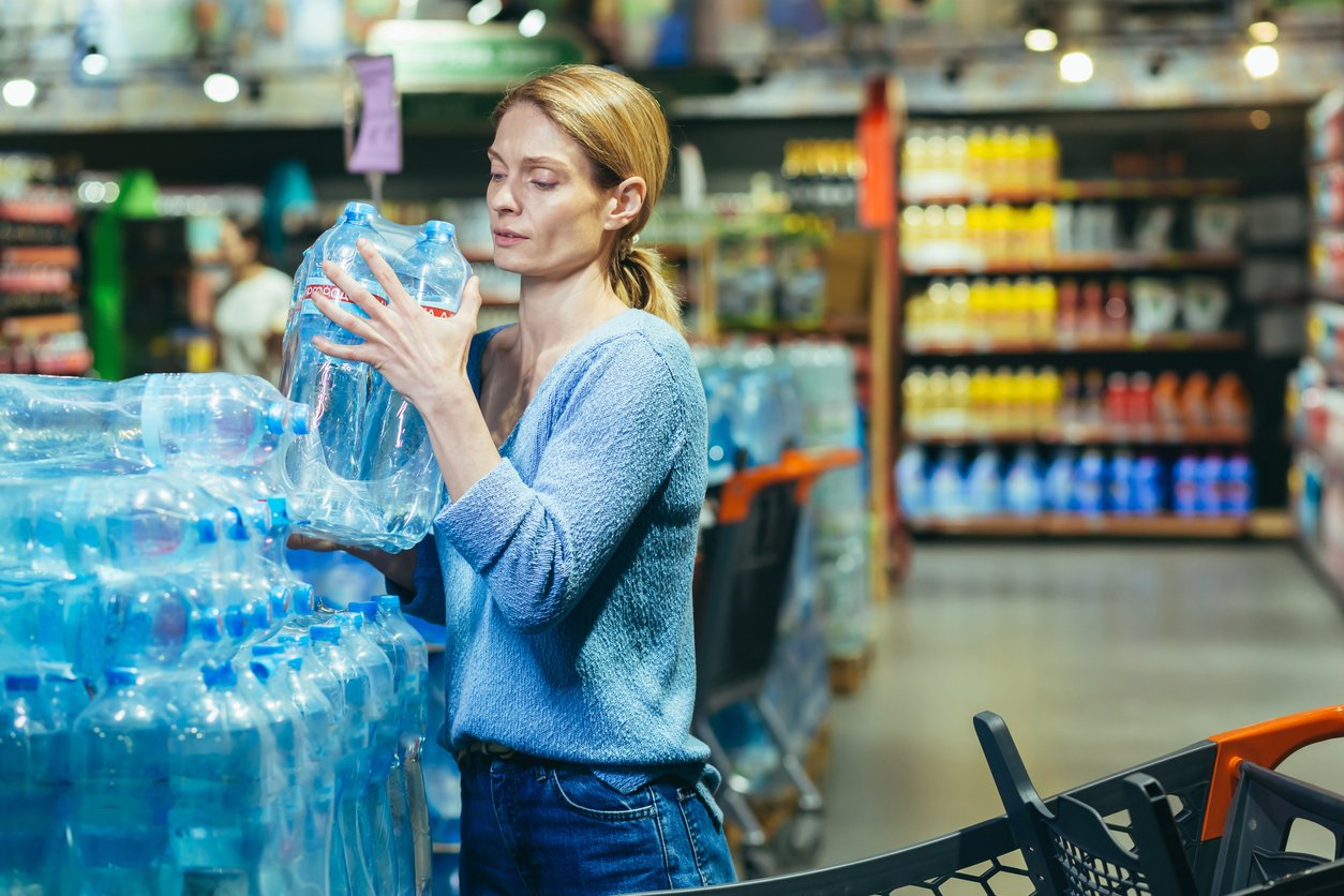 Grocery store, woman lifts water bottles, foreground aisle view, daytime fluorescent light, documentary style, people present.