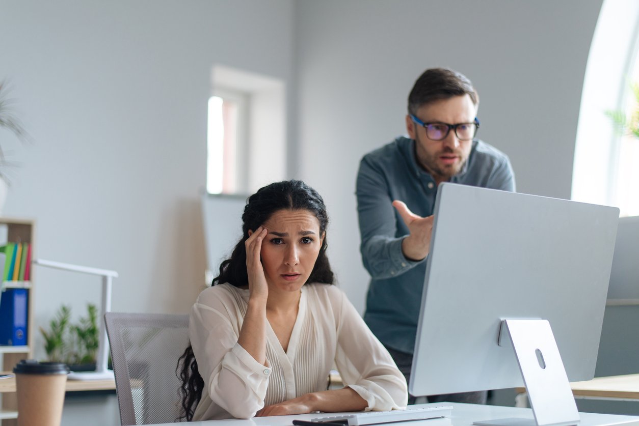 Modern office, stressed woman at desk foreground, man gesturing at computer, bright lighting, documentary style, two people.