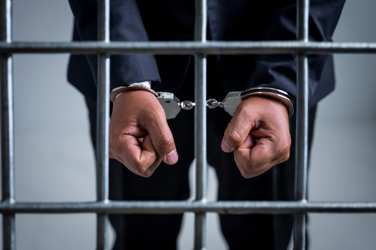 Jail cell interior, cuffed hands gripping bars, upper body cropped, dim jail lighting, documentary style, one suited person.
