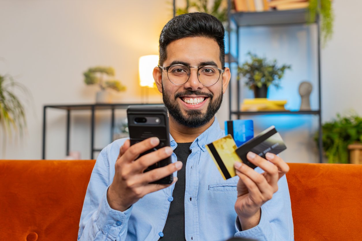 Living room, man with glasses on orange couch, smartphone and credit cards, daylight, editorial travel photo, one person.