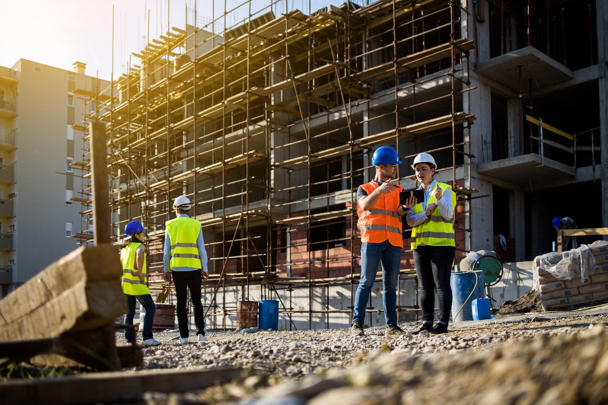 Building site, clipboard review, scaffolding and structure mid-build, daylight, documentary style, several workers present.
