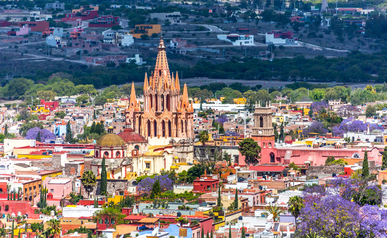 San Miguel de Allende, Parroquia pink spires, vibrant buildings and jacarandas, wide view, sunny day, editorial travel photo, no people.