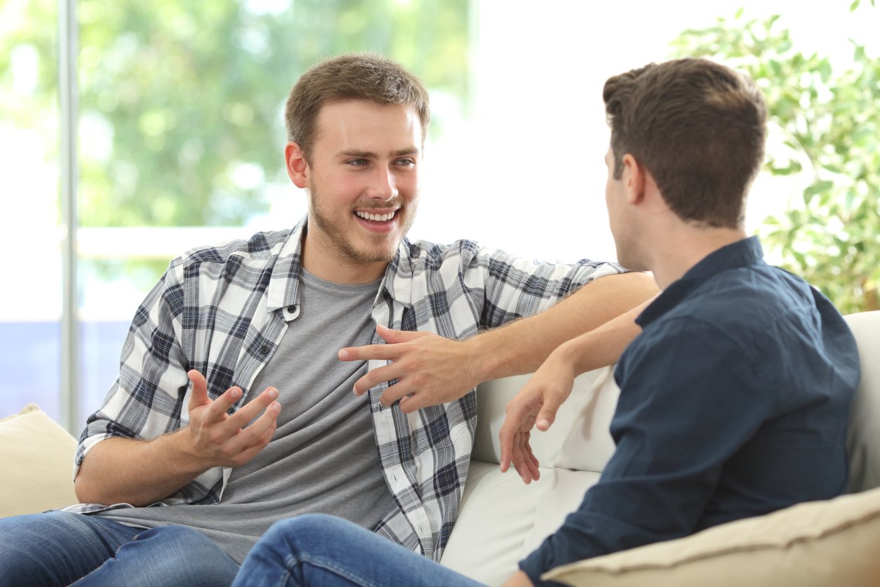 Indoor living room, two men talking on couch, window and green plants behind, bright daylight, documentary style, with people.