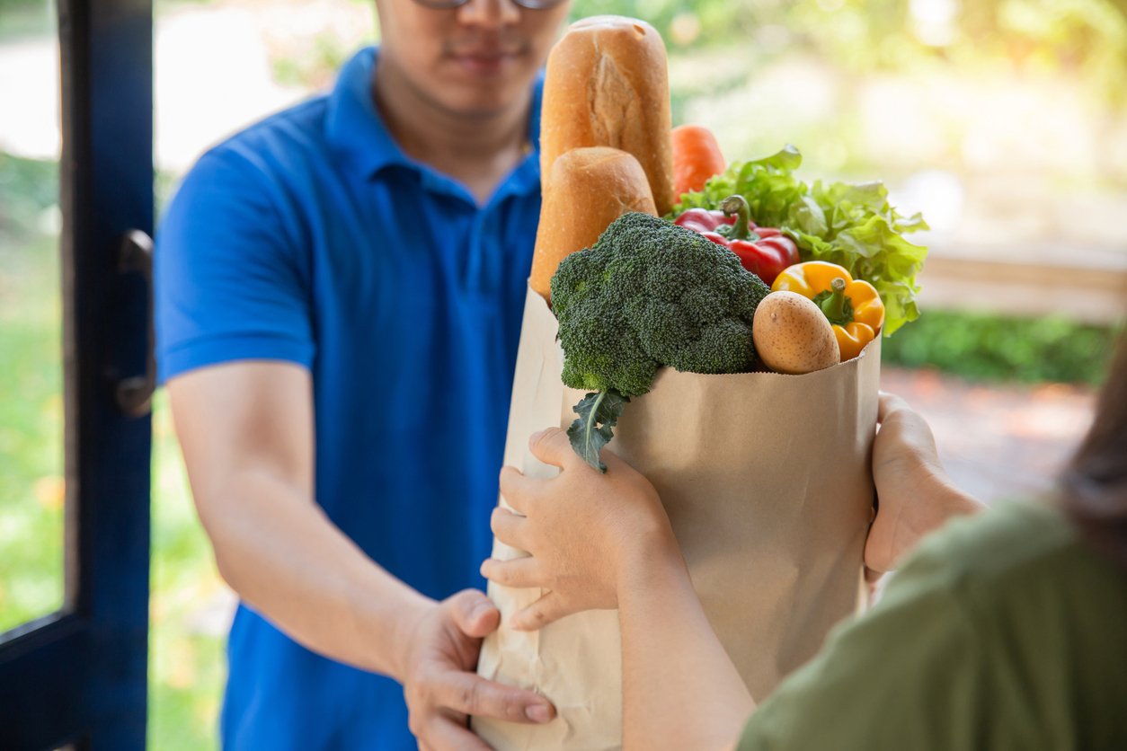 Doorstep, grocery bag exchange, two hands visible foreground, soft daylight, documentary style, people present.