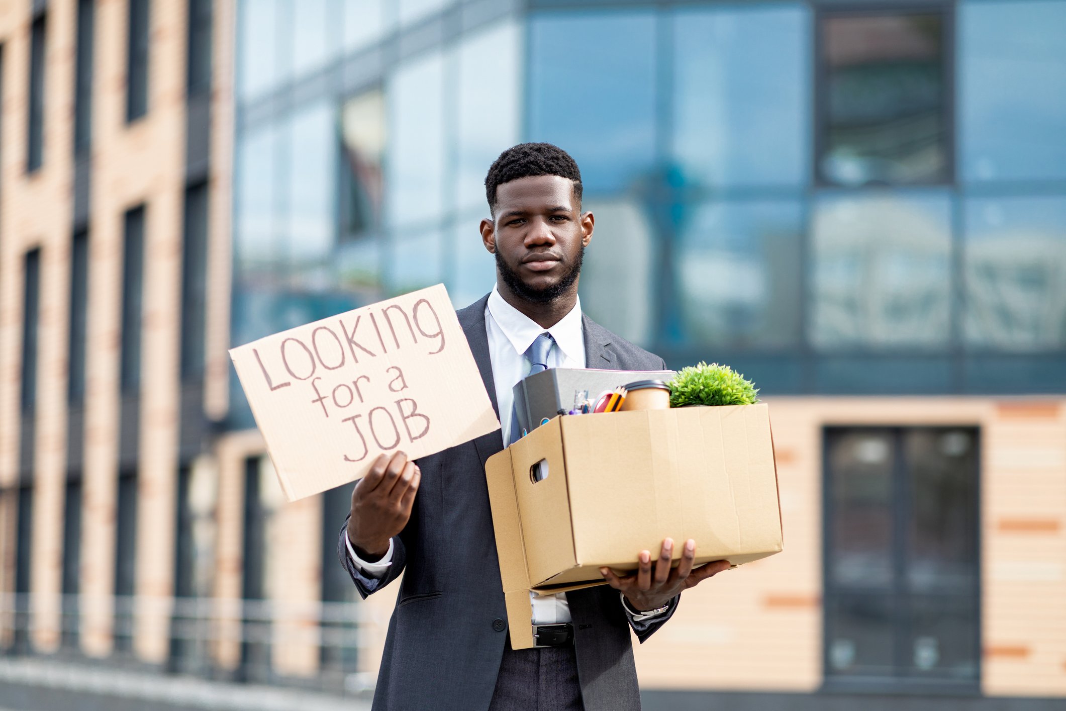 Modern glass building, suited man, holding box and job sign, daylight, editorial travel photo, one person.
