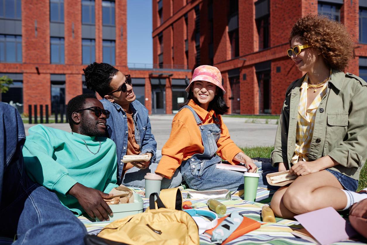 Outdoor lawn, friends sharing picnic food, red brick building backdrop, sunny afternoon light, editorial travel photo, four people.
