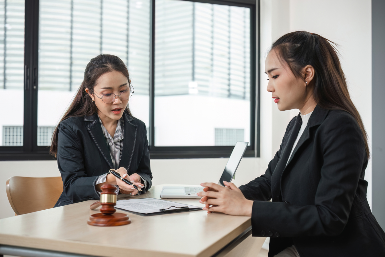 Office conference room, legal documents and gavel, women at desk mid-discussion, bright natural light, documentary style, people.