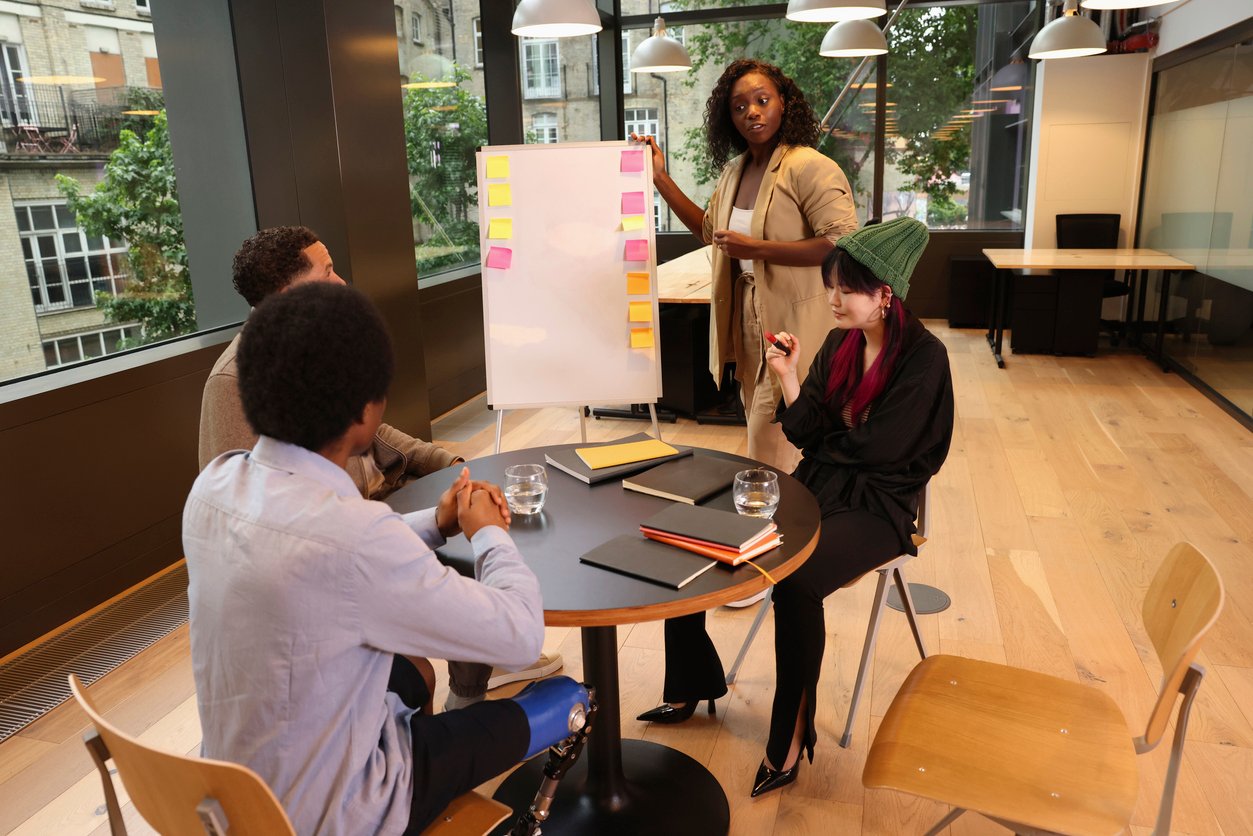 Modern office, whiteboard with sticky notes, presenter leading meeting, daytime natural light, documentary style, four people.