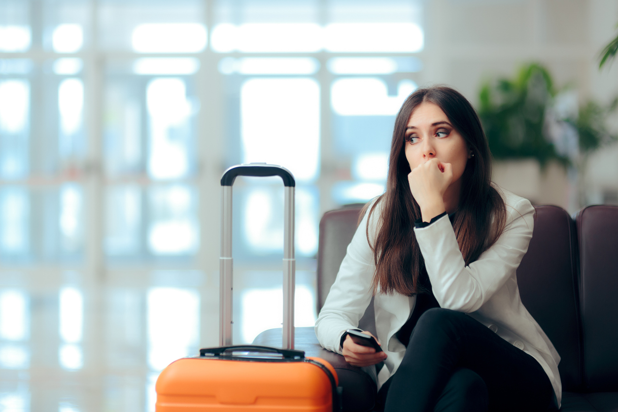 Airport terminal, anxious woman with phone, bench and orange suitcase foreground, daytime bright light, editorial travel photo, one person.
