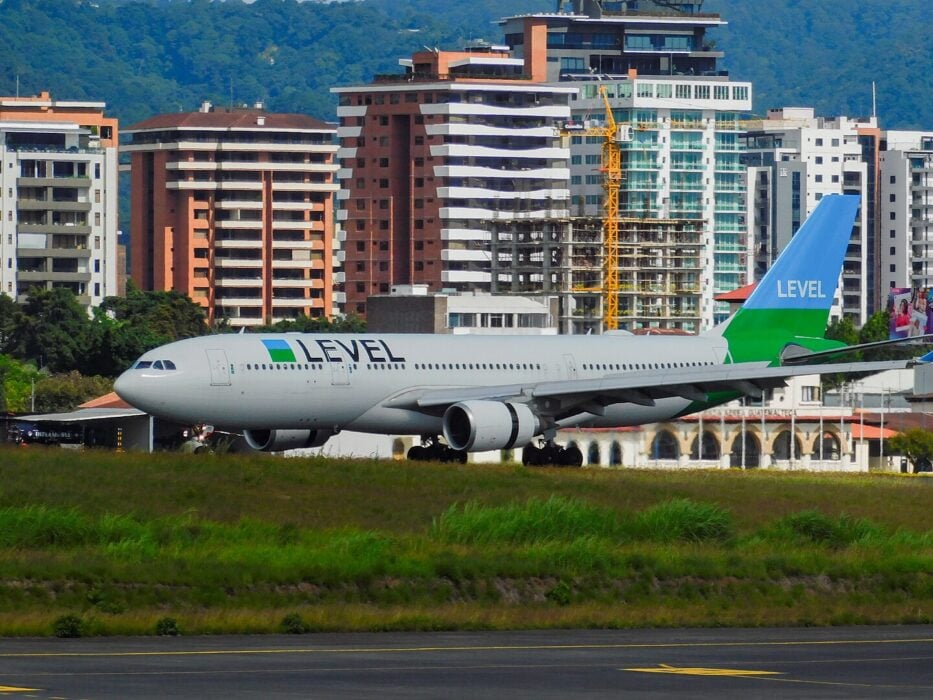 Airport runway, LEVEL airplane taxiing, green grass foreground, modern buildings backdrop, daytime clear light, editorial travel photo, no people.