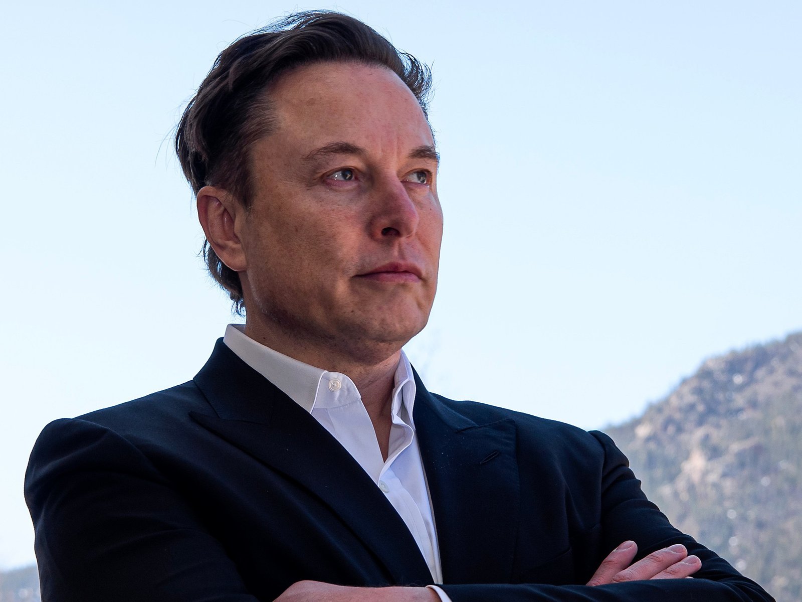 Mountain backdrop, man in black suit foreground, arms crossed center, bright daylight, editorial travel photo style, one person.