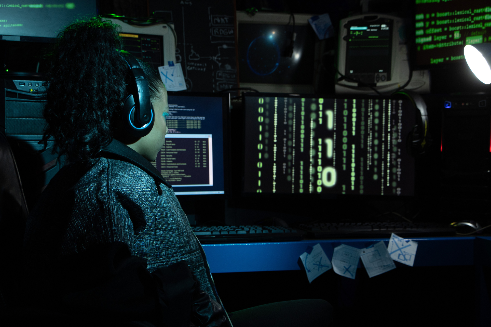 Dark tech room, headphones-wearing person at multi-monitor desk, low light, documentary style, cluttered, one person.