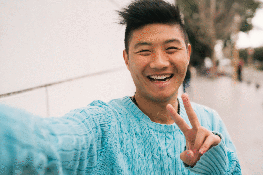 Outdoors near white wall, person smiling with peace sign, selfie composition, daylight, editorial travel photo, one person.