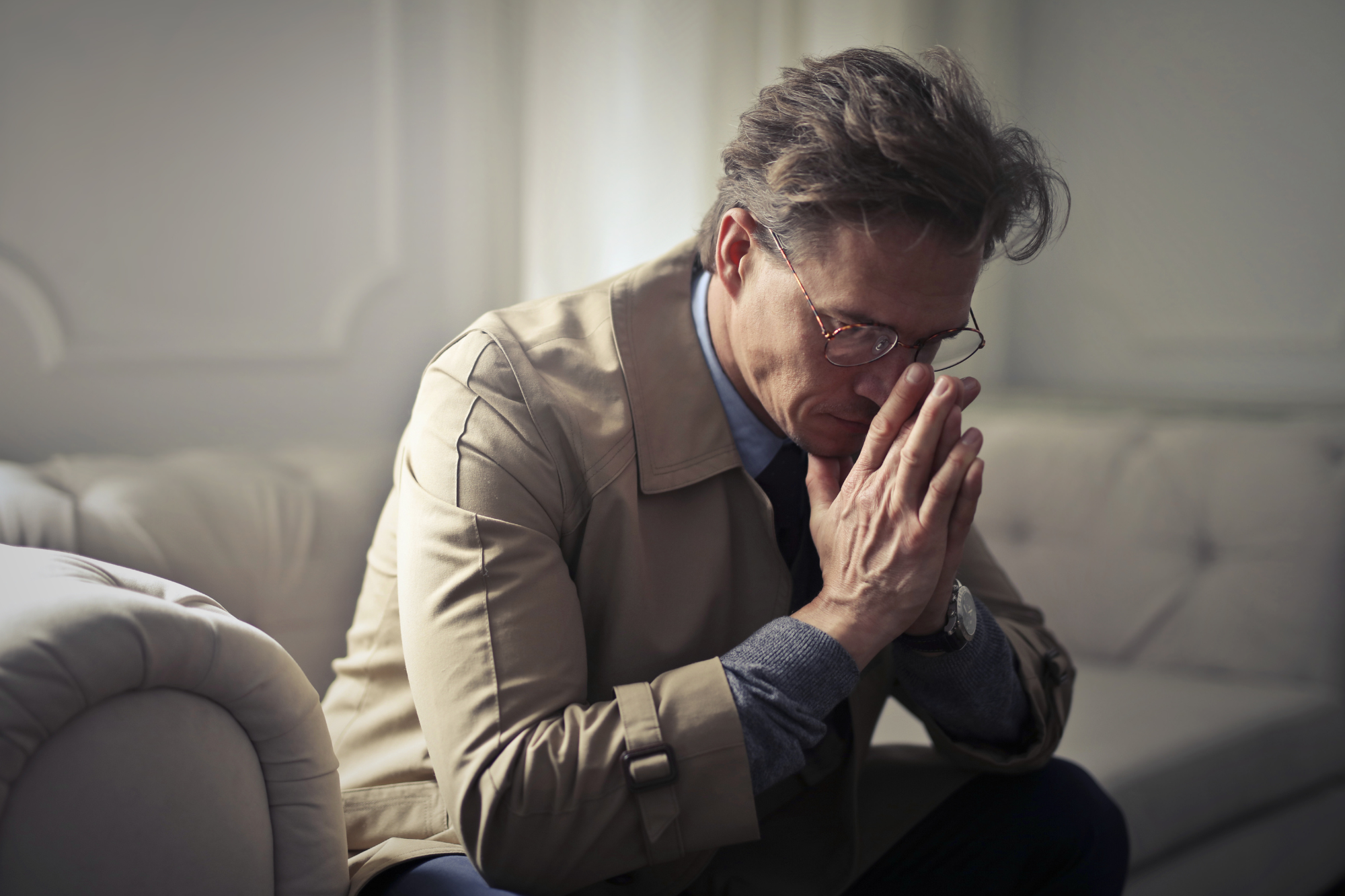 Living room, middle-aged man in glasses and beige coat, hunched forward on couch, soft morning light, documentary style, people.