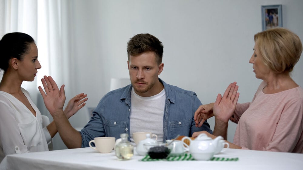 Café table, man flanked by two women arguing, raised hands, daylight indoors, documentary style, people present. mother son girlfriend