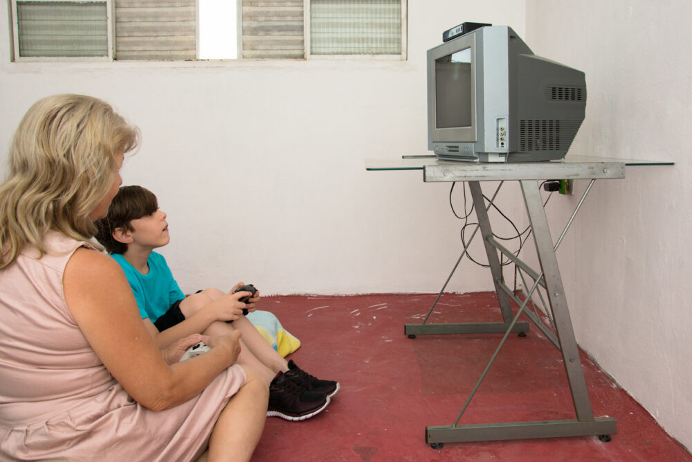 Sparse room with red floor, CRT TV on metal stand, wide shot, daylight, documentary style, woman and boy playing video game.