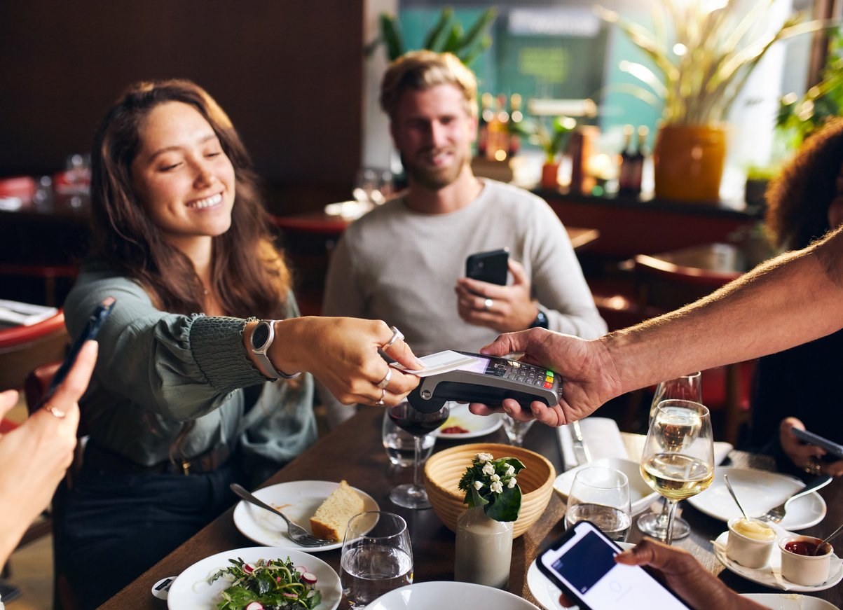 Restaurant table, woman paying with smartphone, server holding card reader, midday sun, editorial travel photo, several people.