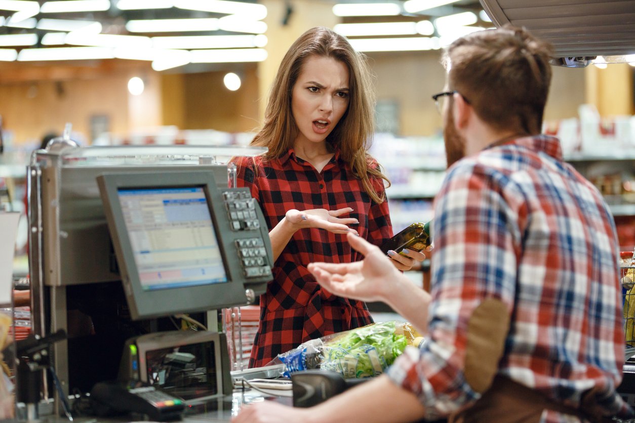 Grocery store checkout, upset woman gesturing at cashier, midday fluorescent lighting, documentary style, groceries visible, people.