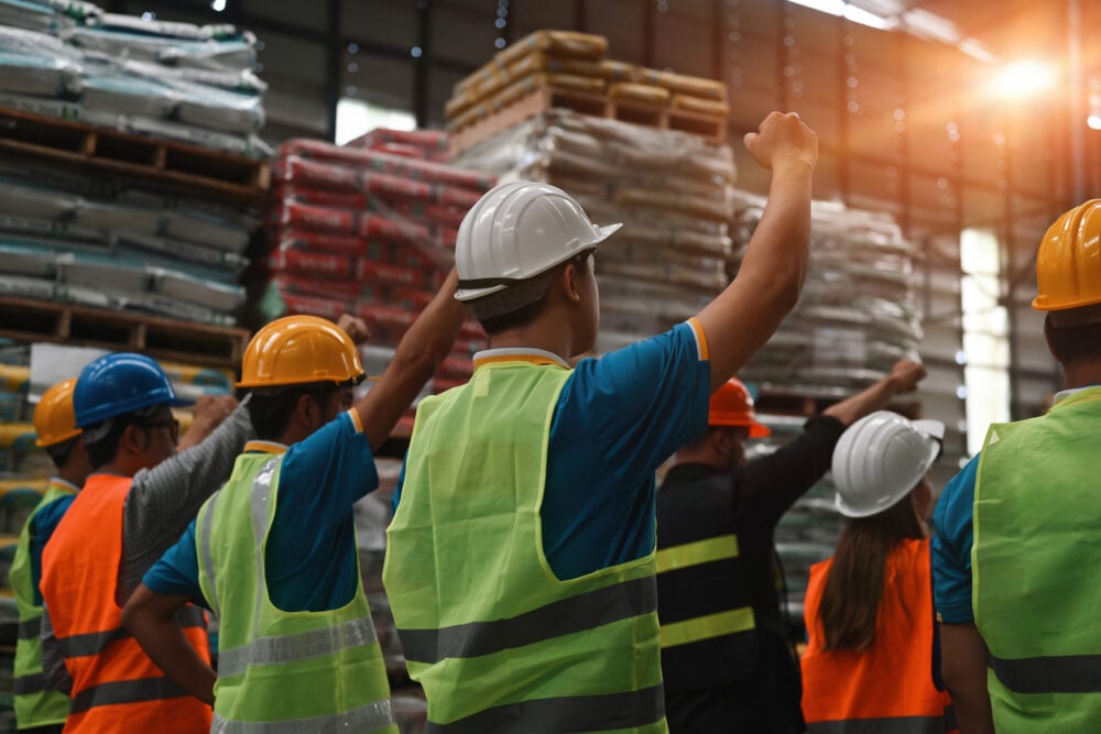 Industrial warehouse, workers united, fists raised foreground, midday sunlight beams in, documentary style, group of people.
