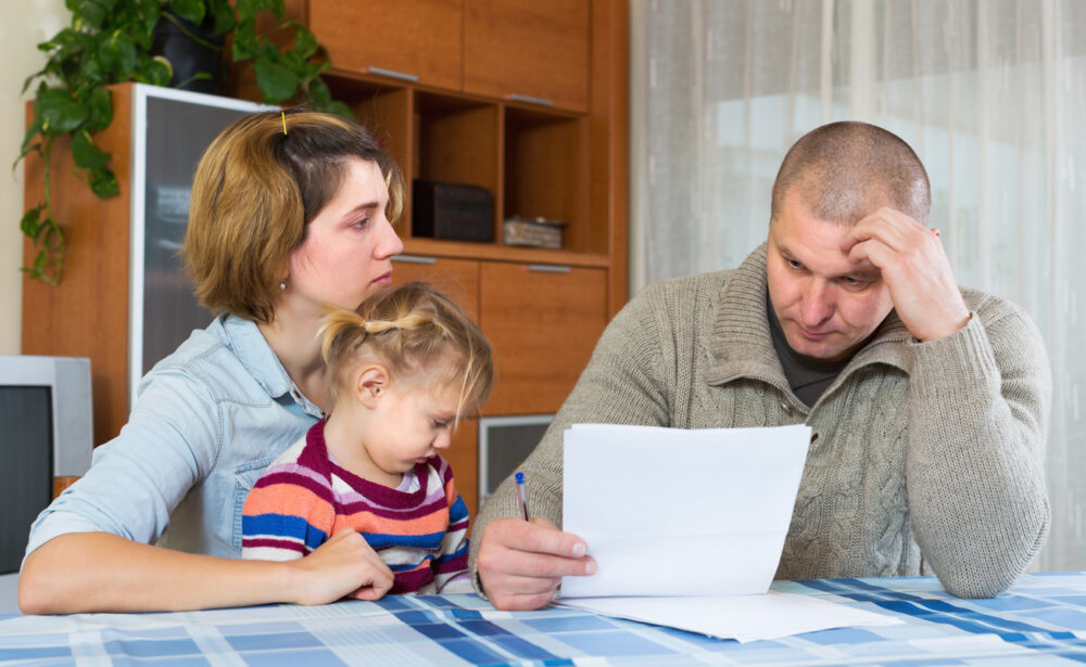 Kitchen table, man anxiously studying documents foreground, soft daylight, documentary style, two concerned family members.