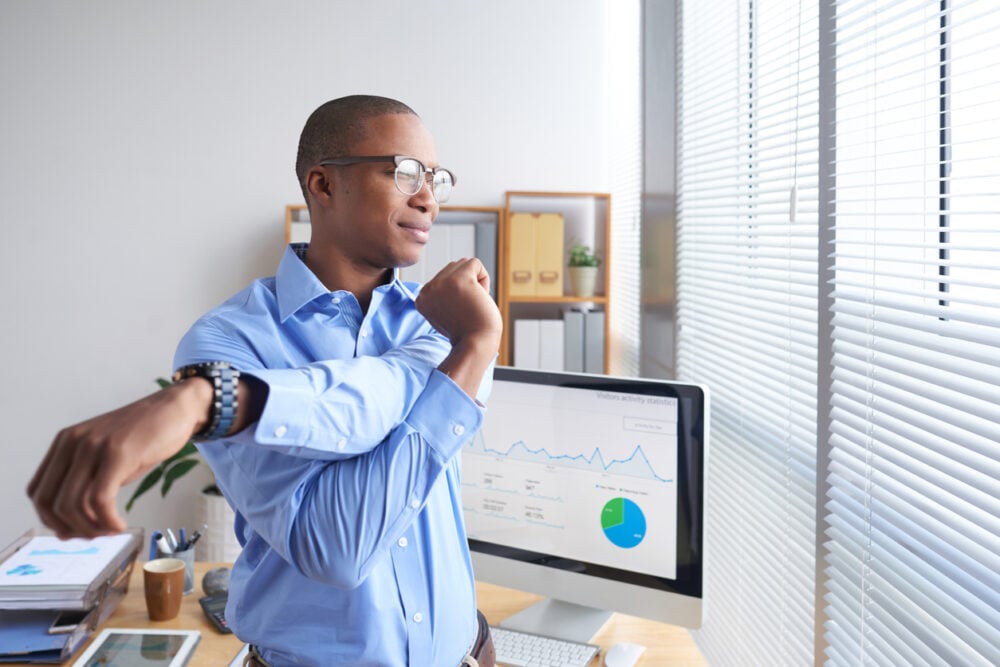 Modern office, man in glasses stretches by desk, computer with graphs visible, morning sunlight through blinds, editorial travel photo, one person.