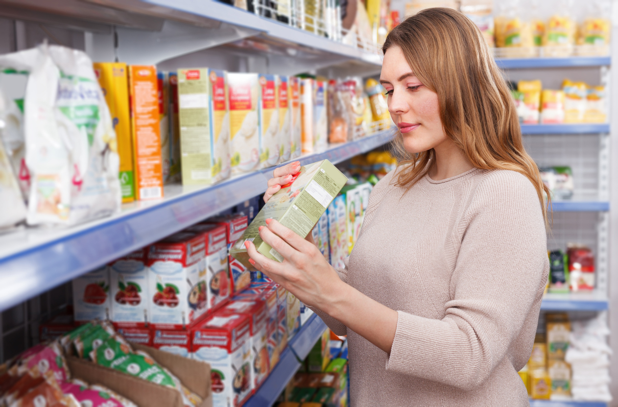 Grocery store aisle, woman holding green package, front view, bright indoor lighting, editorial travel photo, one person.