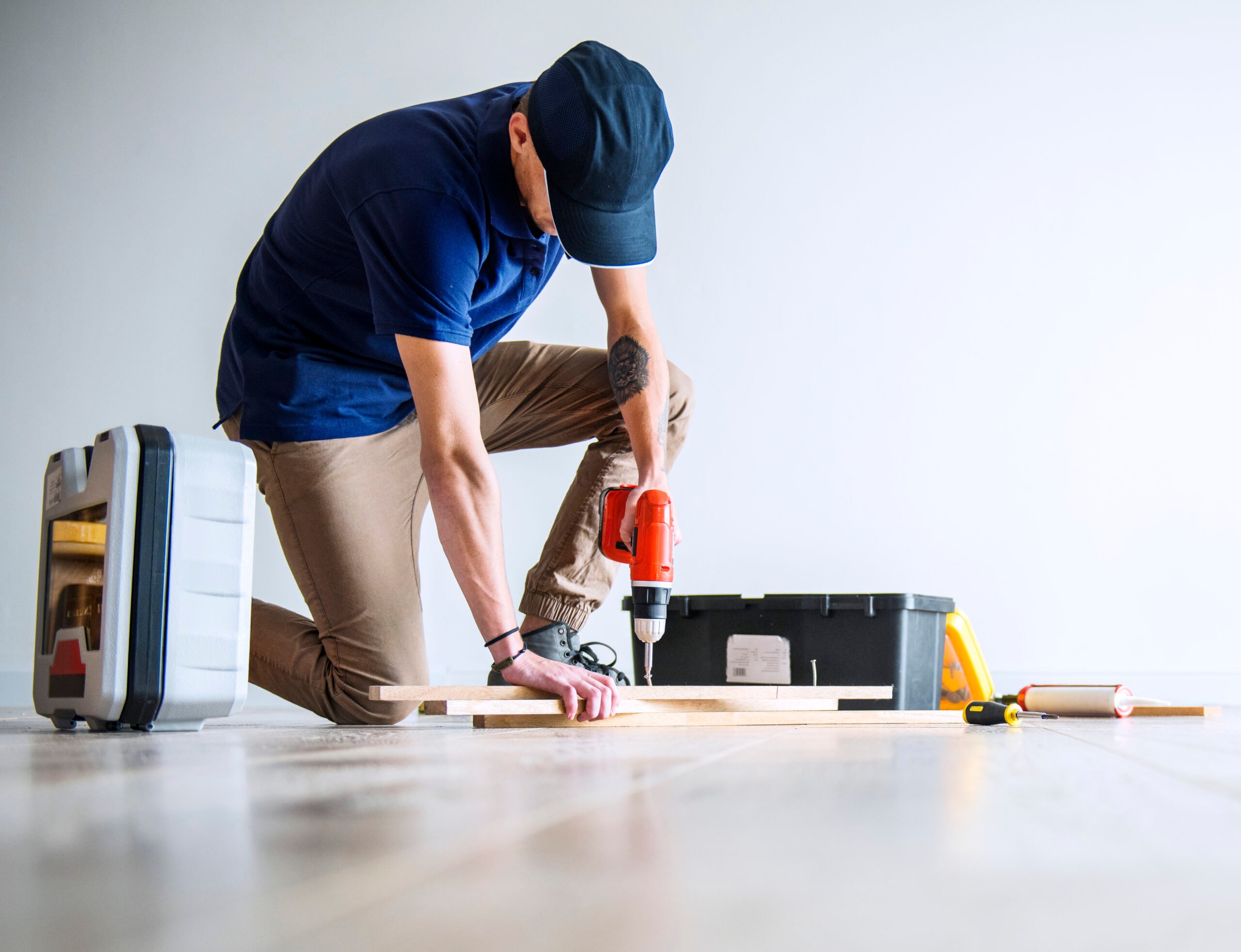 Plain-walled room, worker with drill and tools, kneeling on floor, daytime even light, documentary style, one person.