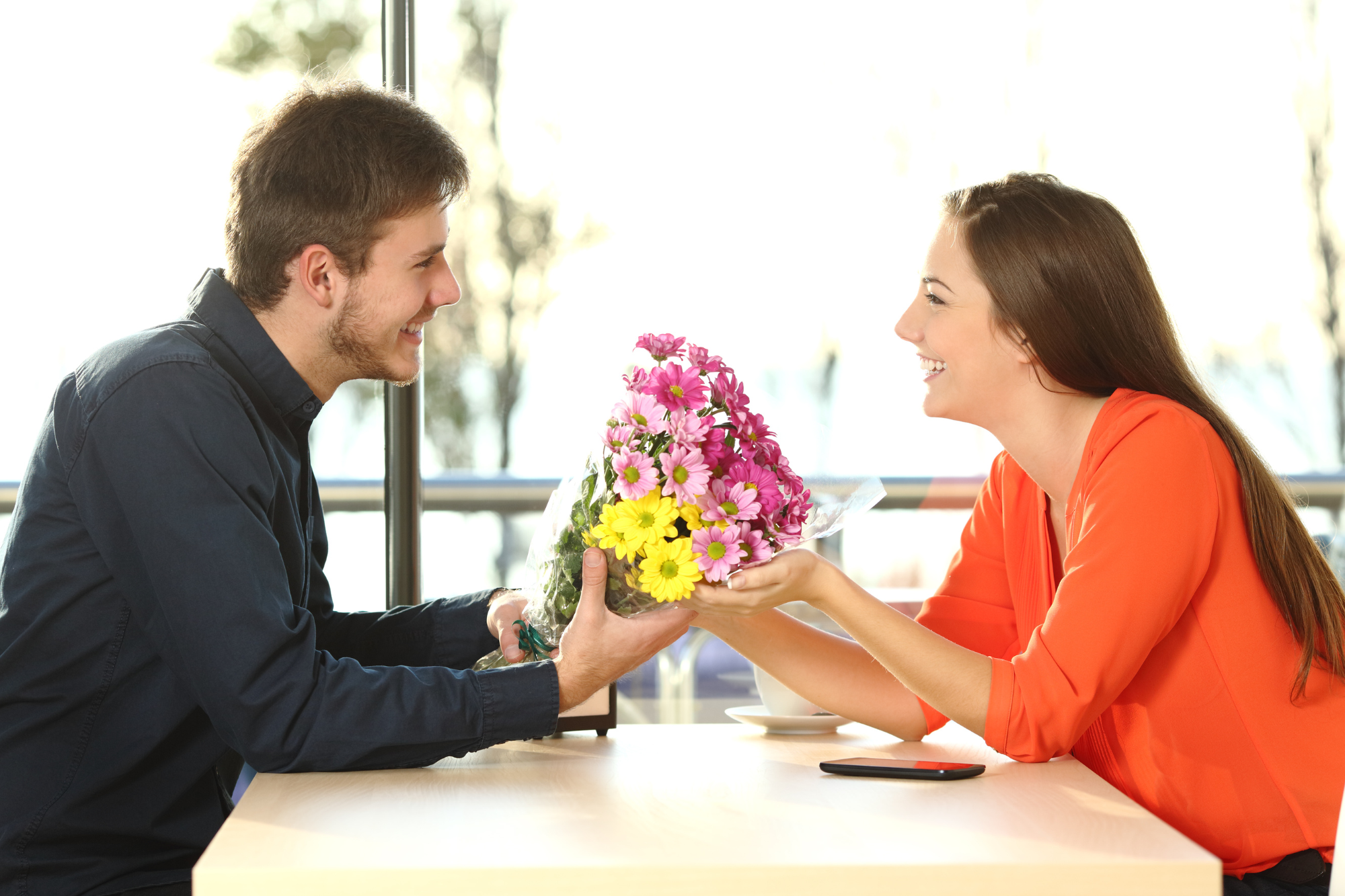 Bright, modern café; man handing bouquet to woman; table centered; daylight streaming in; editorial travel photo; smiling people.