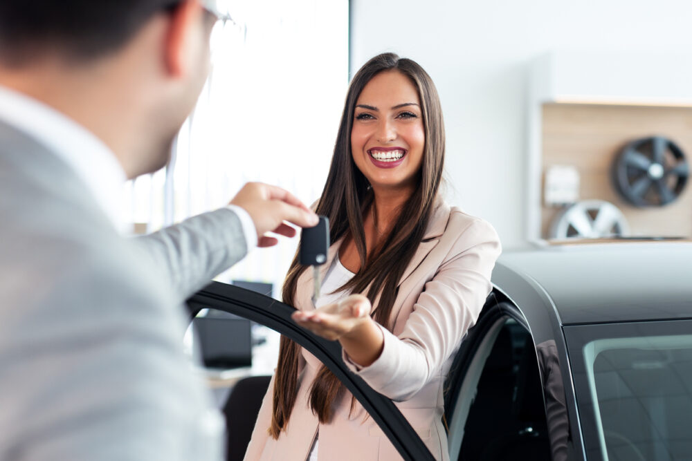 Car dealership, woman receiving keys, open car door foreground, daytime natural light, editorial travel photo, two people.