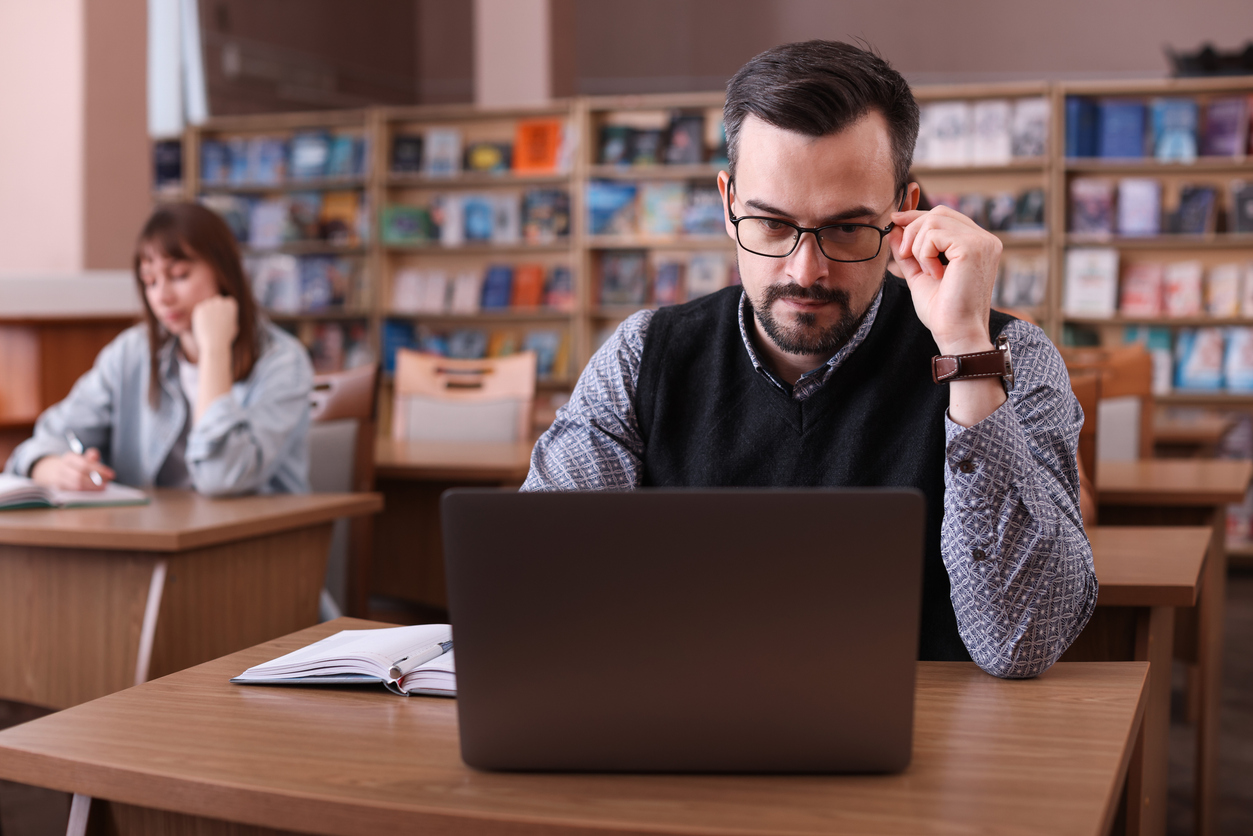 Library, man with glasses at desk, laptop and open notebook, daytime diffused light, editorial travel photo, two people.
