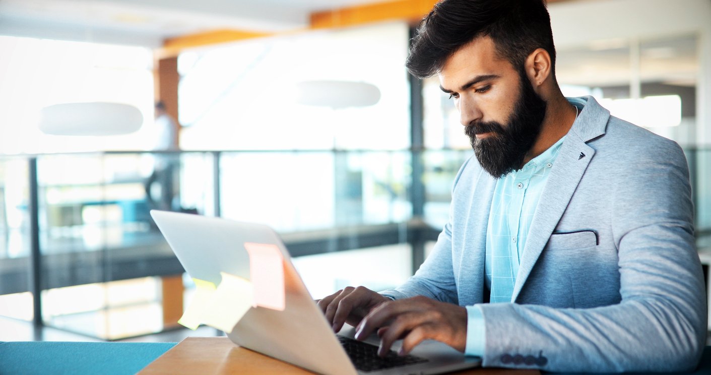 Modern office, bearded man at laptop, sitting at desk, bright natural light, editorial style photo, one person.