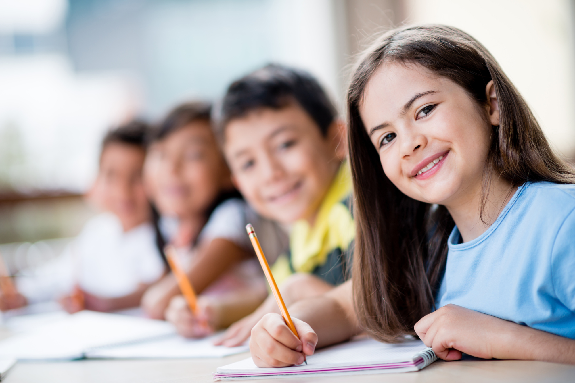 Classroom, girl in blue shirt foreground, other kids blurred behind, daytime natural light, documentary style, people.