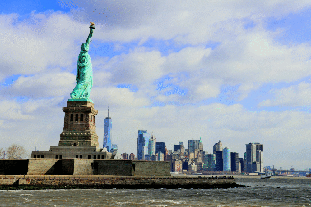 Liberty Island, Statue foreground, NYC skyline behind, daytime partly cloudy, editorial travel photo, no people.