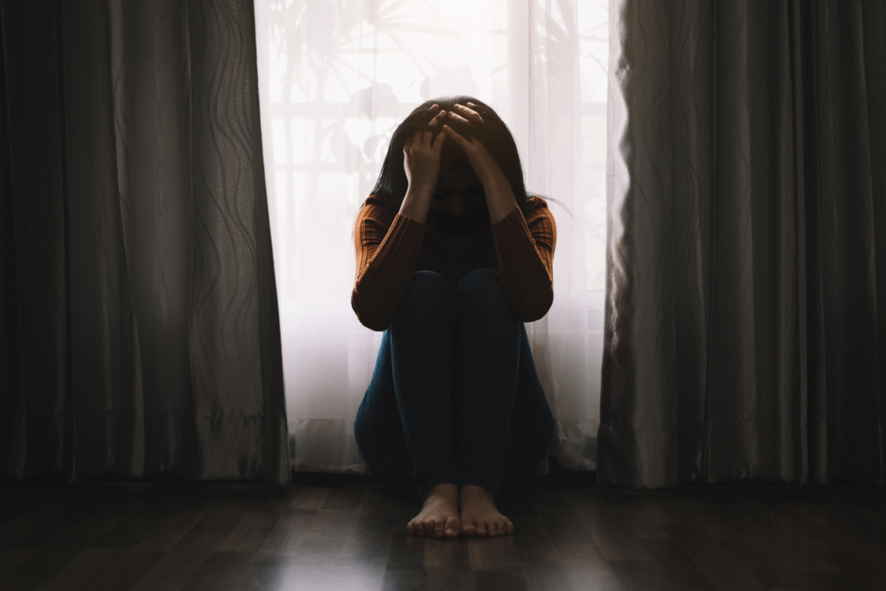 Dimly lit room, window with curtains, person on floor head in hands, morning light streaming, documentary style, one person.