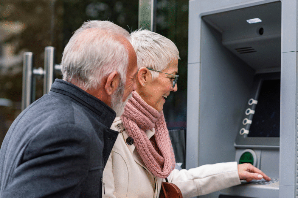Outdoor ATM, elderly woman using machine, man beside her, daytime natural light, editorial travel photo, two people.