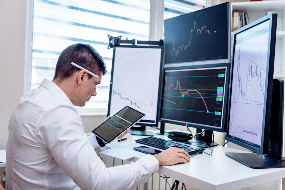 Modern office, man at desk with multiple screens, midday light, financial charts clear, documentary style, one focused person.