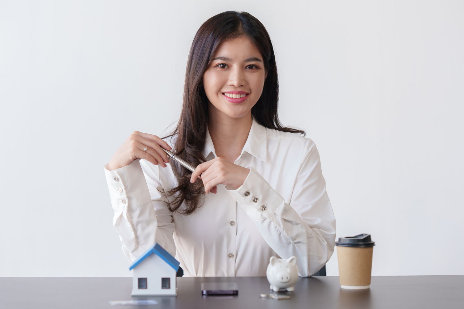 Indoor office, woman with house model and piggy bank at table, daytime natural light, editorial travel photo, one person.