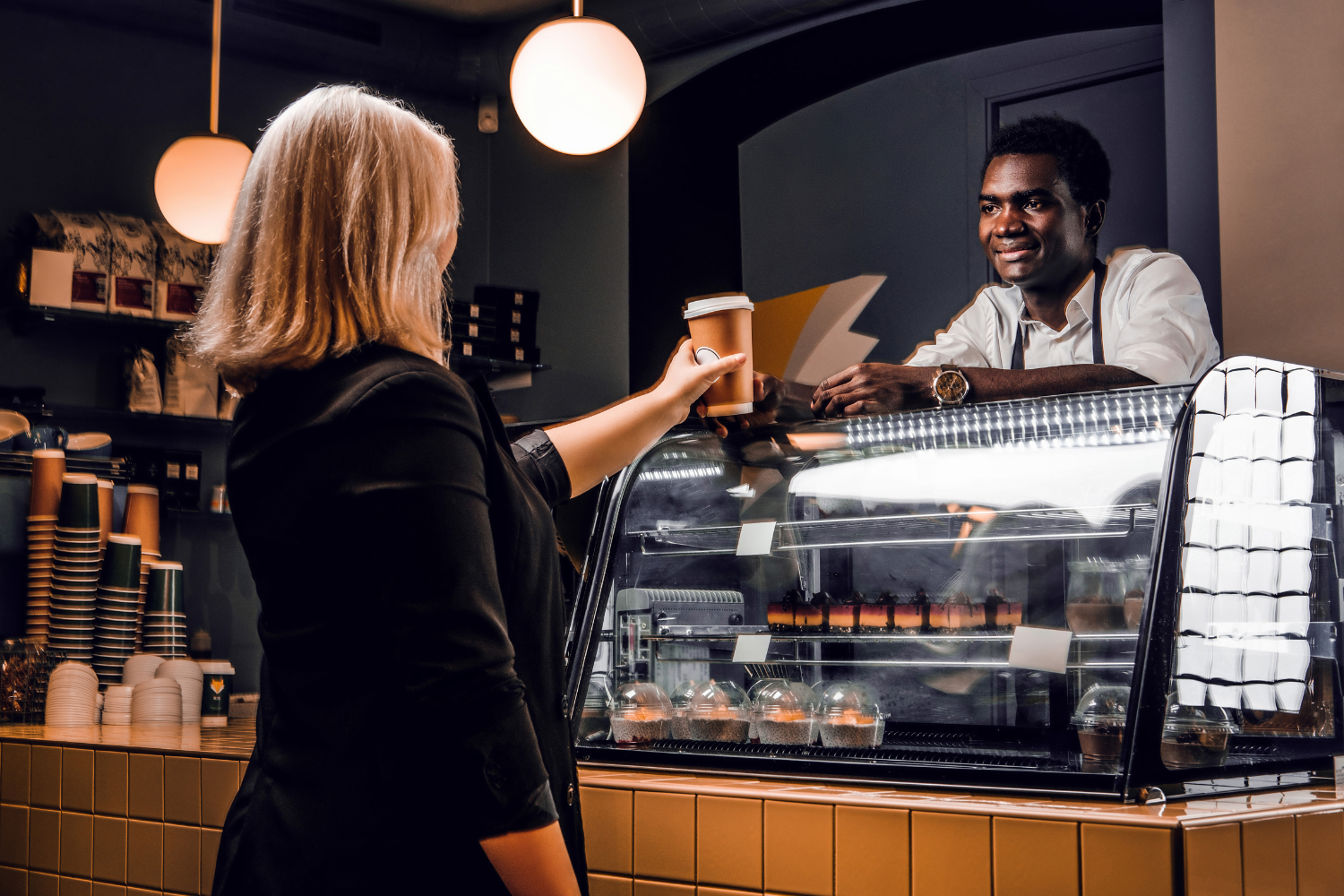 Café counter, barista handing coffee to blonde woman, glass pastry case foreground, warm lighting evening, editorial travel photo, two people.