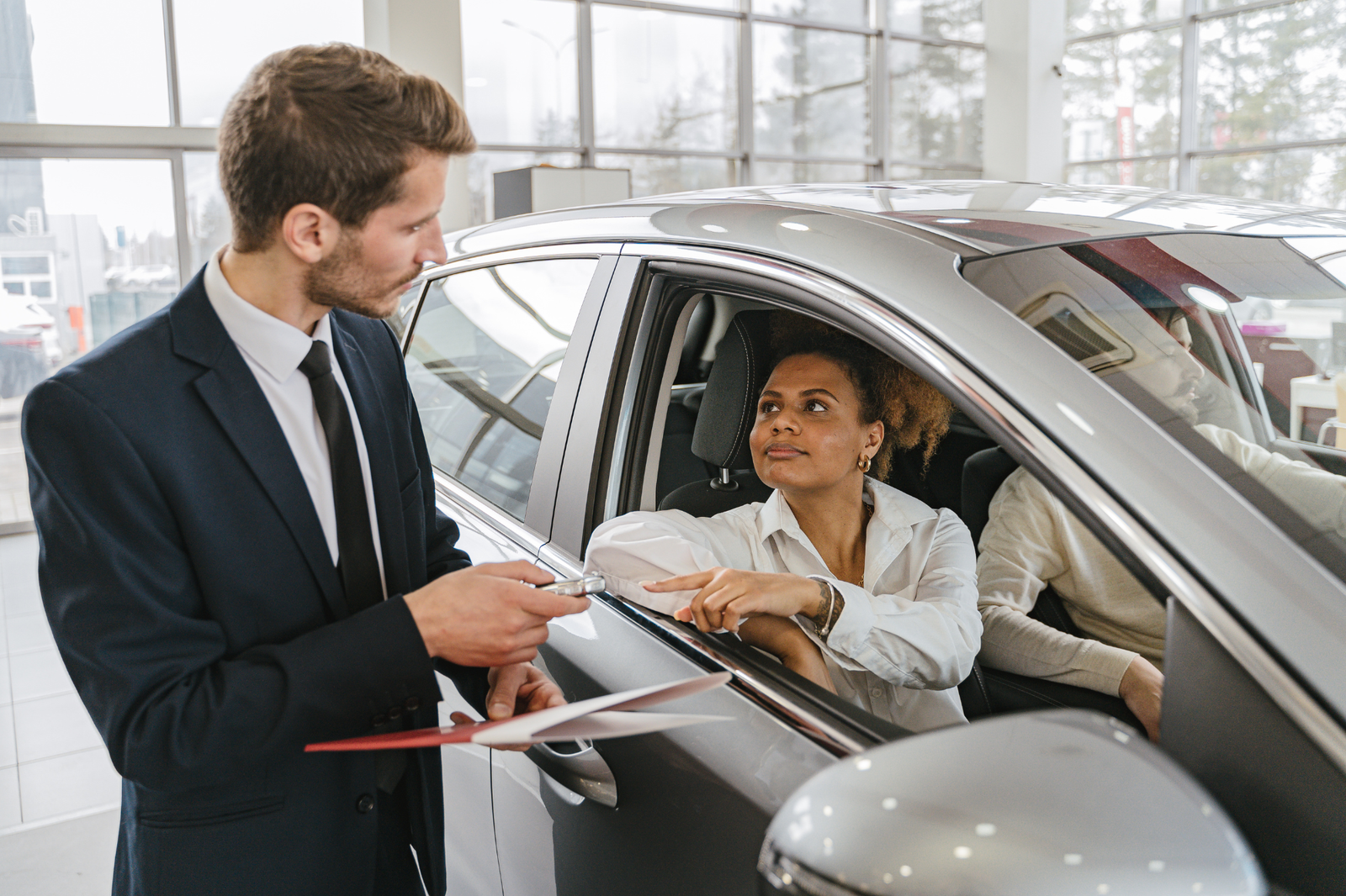 Dealership interior, silver car foreground, salesperson showing key, daytime natural light, documentary style photo, three people.