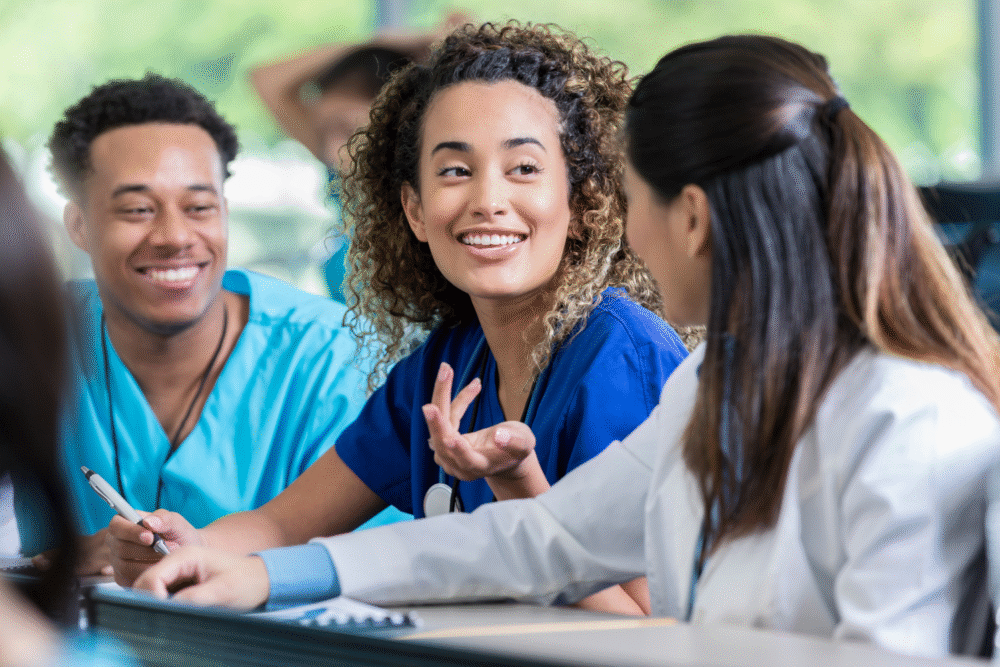 Bright classroom, medical students at table, casual group shot, daytime sunlit, documentary style, three smiling people.