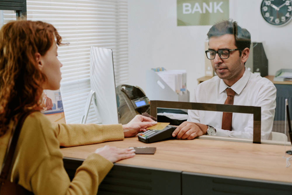 Bank counter, woman in brown jacket paying by card, teller behind glass, bright daylight, documentary style, two people.