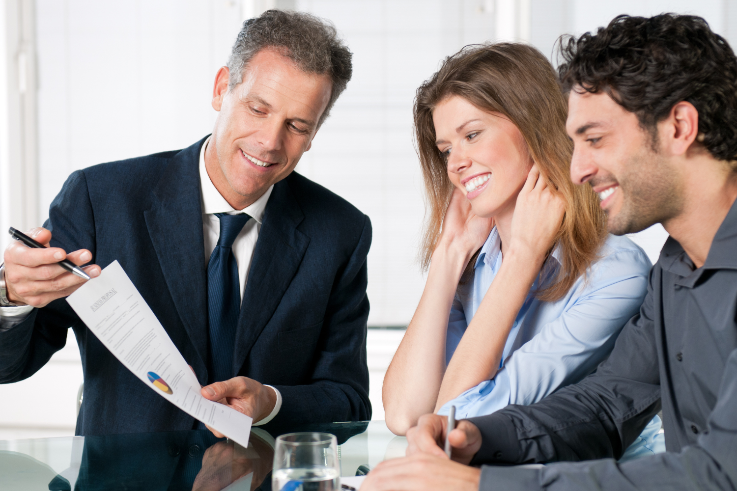 Office interior, suited man at desk with young couple, eye-level view, daytime, documentary style, 3 people present.
