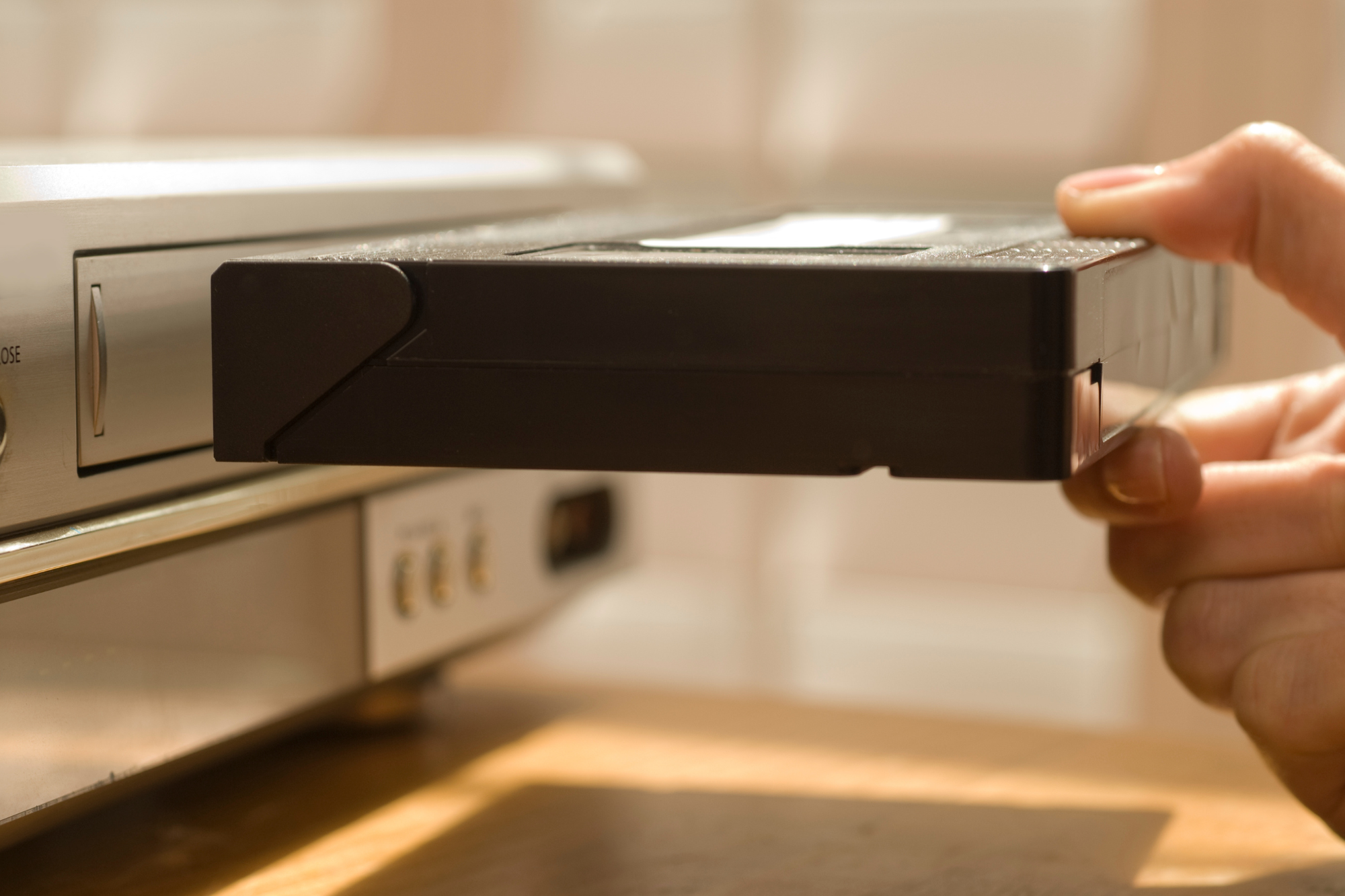 Living room, VHS tape entering silver VCR, close-up with sunlit detail, morning light, documentary style, no people.