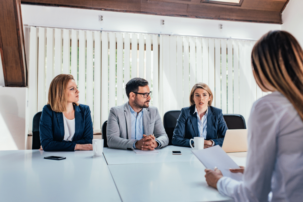 Conference room, three professionals at table, focus on interviewer’s back, natural daylight, documentary style, with people.