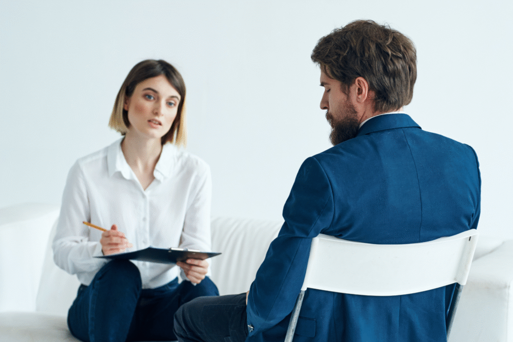 Minimalistic office, woman with clipboard foreground, man seated beside her, daylight, documentary style, two people.